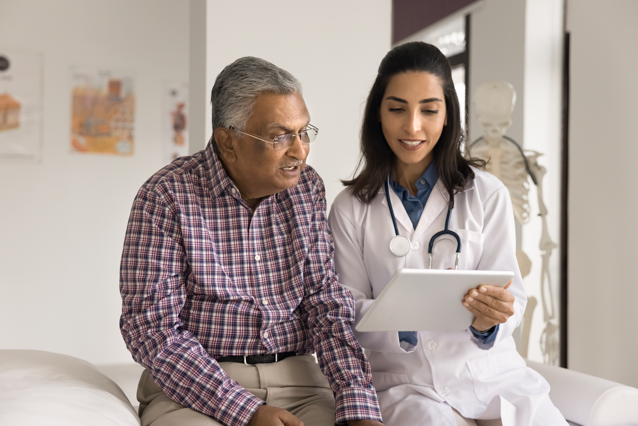 Positive young Indian doctor woman showing electronic content on tablet computer to senior Indian man, speaking to patient, explaining healthcare examination results